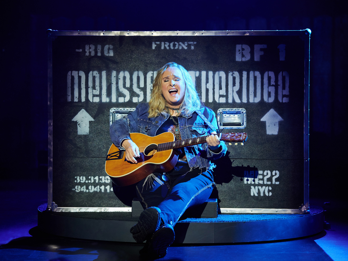 Melissa Etheridge sits against a trunk playing guitar