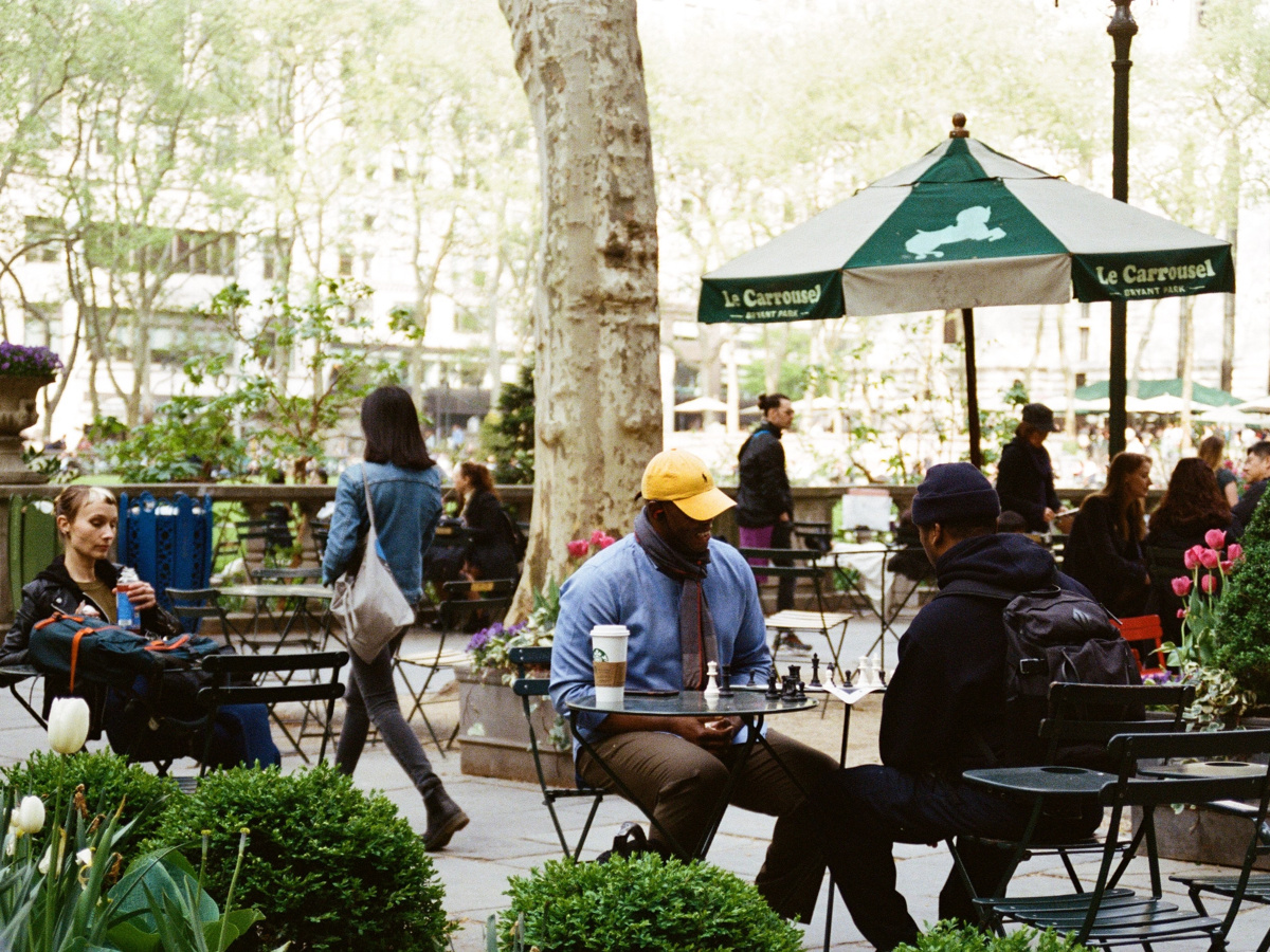 View of Bryant Park. People playing chess and people walking through the park.
