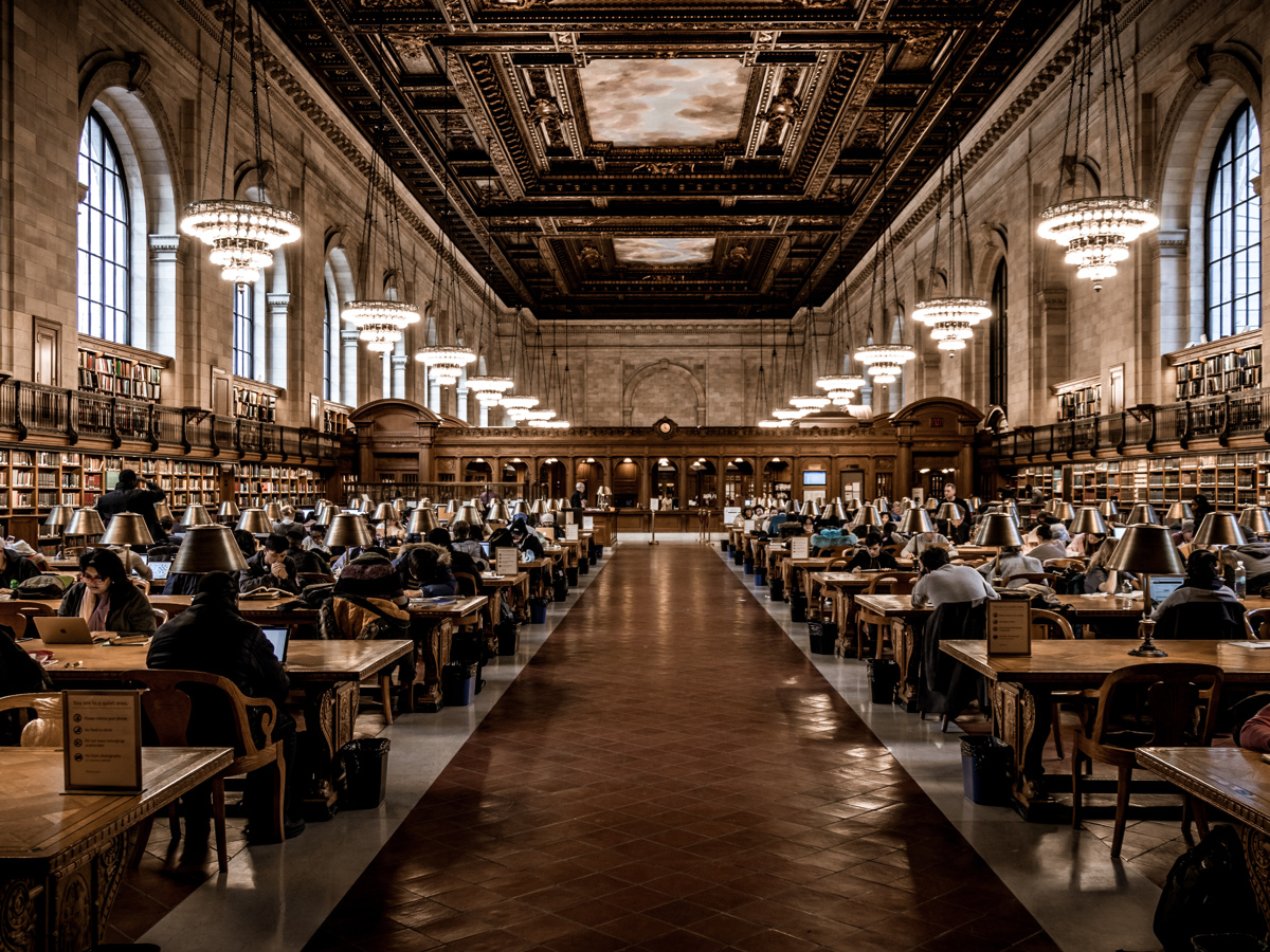 Inside the NY Public Library. People sitting, reading at large tables with high, ornate ceiling above.