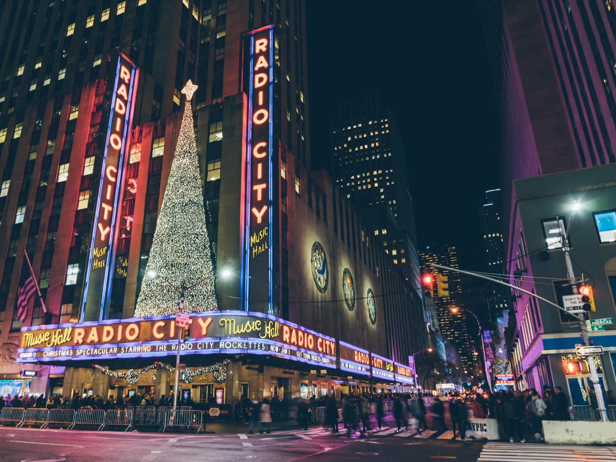 Radio City Music Hall at night, in the holiday season. The venue's lights are illuminated and there is an addition of a Christmas tree.